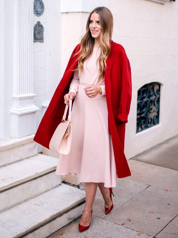 Woman in a blush pink dress with a red coat, red heels and carrying a red bag, standing on a sidewalk.