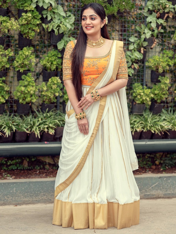 Actress Arya Babu in a cream saree with orange blouse and gold trim, adorned with gold necklace and bangles. Actress Arya Babu in a cream saree with orange blouse and gold trim, adorned with gold necklace and bangles.