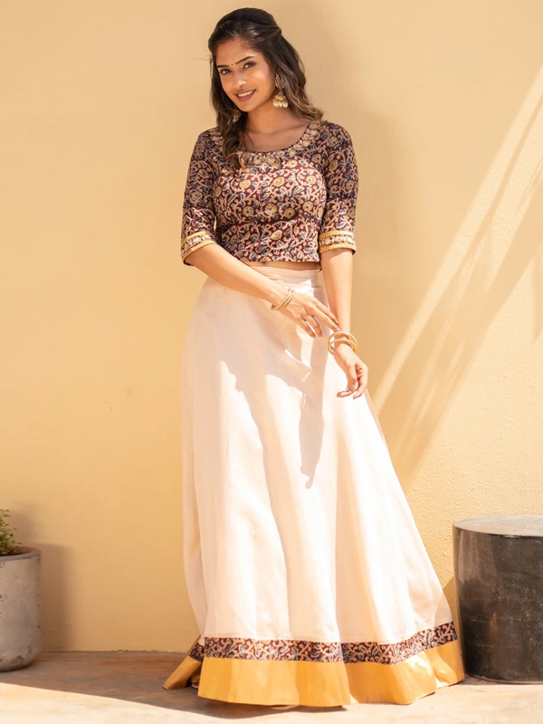 Woman wearing a cream lehenga with a brown Kalamkari blouse and yellow hem detailing. Woman wearing a cream lehenga with a brown Kalamkari blouse and yellow hem detailing.