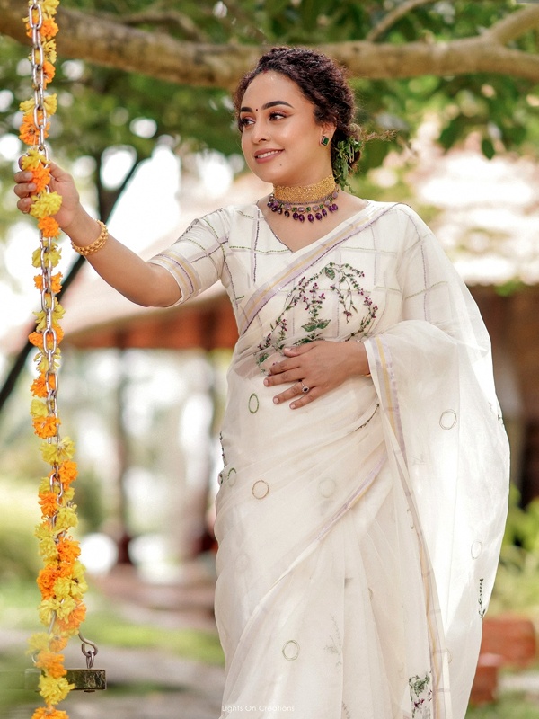 Actress Pearle Maaney in a white floral saree with silver accents, holding a flower garland. Actress Pearle Maaney in a white floral saree with silver accents, holding a flower garland.