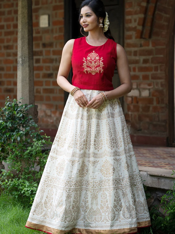 Woman wearing a red top with floral embroidery and an off-white skirt with traditional designs, complemented by gold bangles. Woman wearing a red top with floral embroidery and an off-white skirt with traditional designs, complemented by gold bangles.