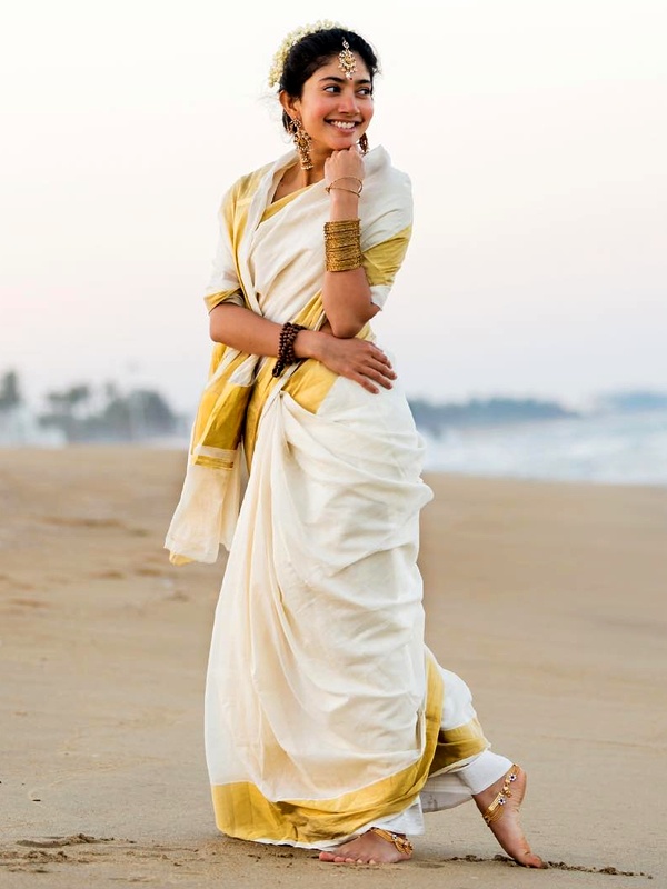 Actress Sai Pallavi on the beach, wearing a white saree with golden border and gold bangles, earrings and a necklace. Actress Sai Pallavi on the beach, wearing a white saree with golden border and gold bangles, earrings and a necklace.
