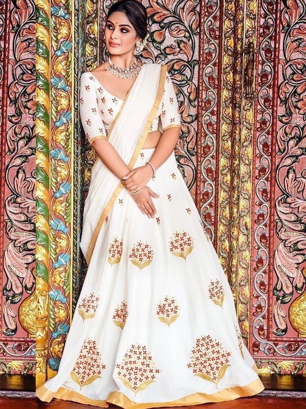 Actress Samyuktha in a white lehenga with gold and red floral embroidery, standing against a vibrant backdrop. Actress Samyuktha in a white lehenga with gold and red floral embroidery, standing against a vibrant backdrop.