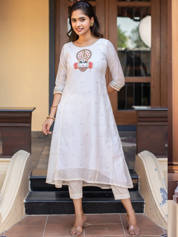 Woman in a white asymmetric kurta with a central Kathakali motif and embellished cuffs, paired with white pants. Woman in a white asymmetric kurta with a central Kathakali motif and embellished cuffs, paired with white pants.