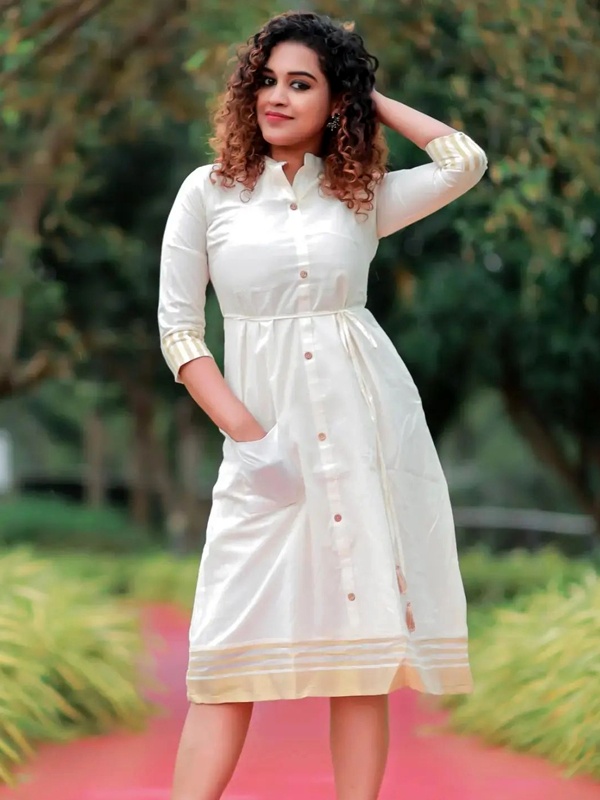 Woman in a white dress with Kasavu border detailing at the hem and cuffs, standing outdoors. Woman in a white dress with Kasavu border detailing at the hem and cuffs, standing outdoors.