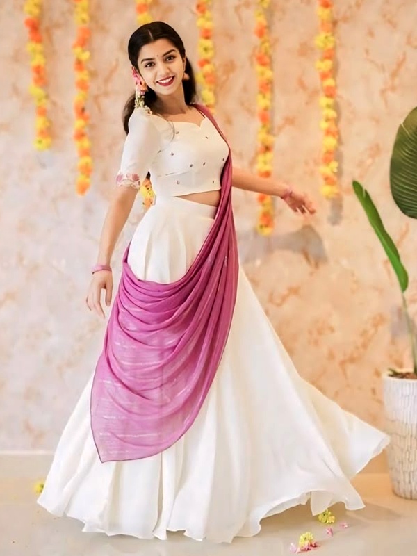 Woman wearing a white lehenga with a pink dupatta, adorned with earrings, standing in front of a floral backdrop. Woman wearing a white lehenga with a pink dupatta, adorned with earrings, standing in front of a floral backdrop.