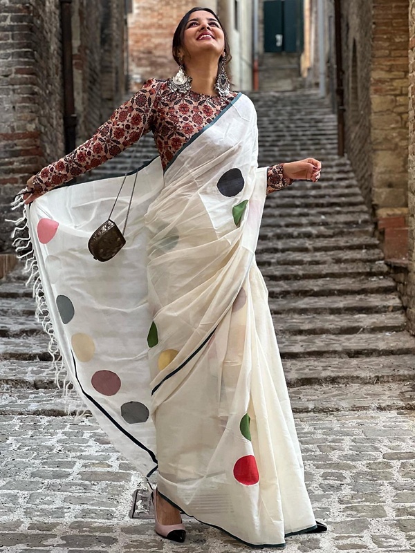 Woman in a white saree with colorful polka dots and a red patterned blouse, accessorized with a small black bag. Woman in a white saree with colorful polka dots and a red patterned blouse, accessorized with a small black bag.