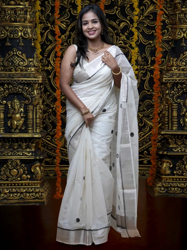 Woman in a white saree featuring a distinctive silver jerry border and black accents, complemented by gold jewellery. Woman in a white saree featuring a distinctive silver jerry border and black accents, complemented by gold jewellery.