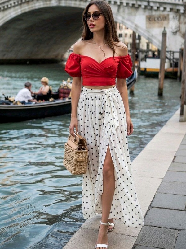 Woman wearing red off-shoulder puff sleeve crop top with sweetheart neckline, white polka dot maxi skirt with high slit, silver sandals and woven handbag